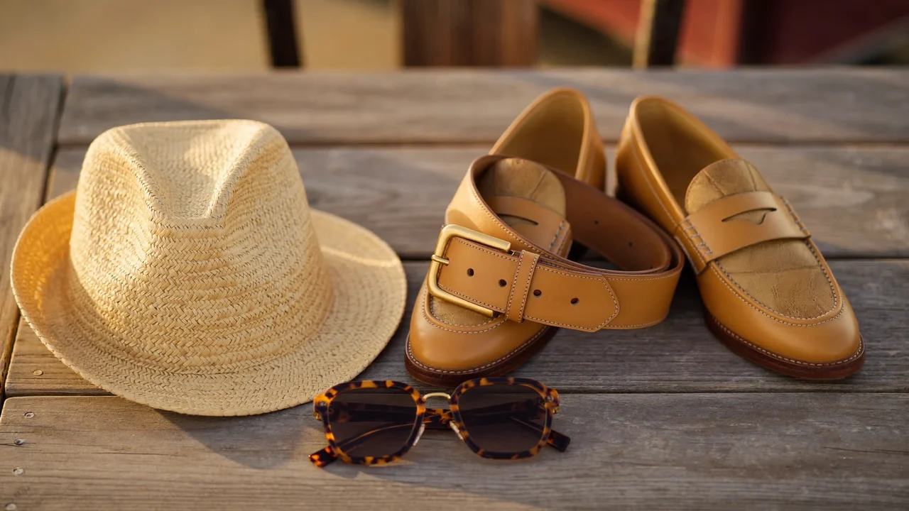 Accessoires homme sur une table en bois : ceinture, lunettes, chapeau de paille, mocassins