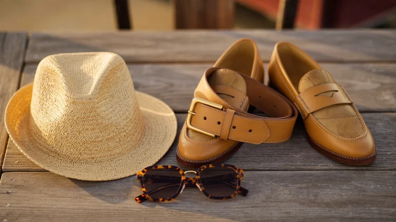 Accessoires homme sur une table en bois : ceinture, lunettes, chapeau de paille, mocassins